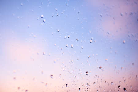 Raindrops on the windshield of the car in the early morning. Transparent glass after rain, pinky abstract photo. Texture of glass with scattered rain drops and a blurry background in the duskの写真素材