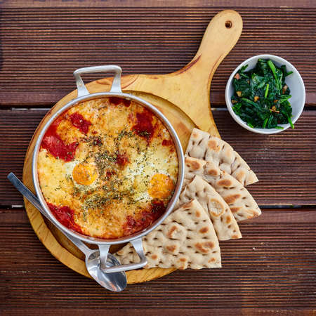 Scrambled eggs with tomatoes, cheese and spices in an aluminum frying pan with handles with homemade thin lavash and juicy greens on a wooden brown table. The photo was taken under natural lightの写真素材