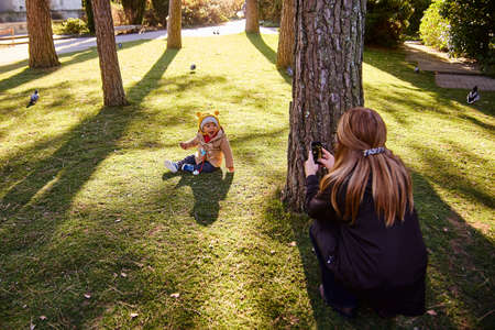 Lisbon, Portugal - January, 2018. Gulbenkian park and garden. Mom and son are playing in the park. A little boy is sitting on the grass, and a woman is taking pictures of him on a mobile phoneのeditorial素材