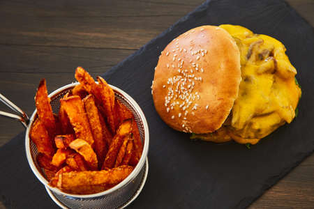 Cheeseburger with a rolled up bun with sesame, melted yellow cheese, green fresh salad and fresh chop on a black board on a dark wooden table. With fried potatoes with large salt in a shiny metal jarの写真素材