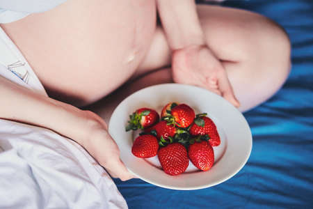 Attractive young pregnant woman sits on a bed and eats a ripe strawberry. Food for a happy pregnant...