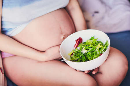 The concept of a healthy diet of pregnancy. A woman in a pregnant state sits on a bed and holds a white bowl with a green and red salad. Healthy eating during pregnancy. Food for pregnant womenの写真素材