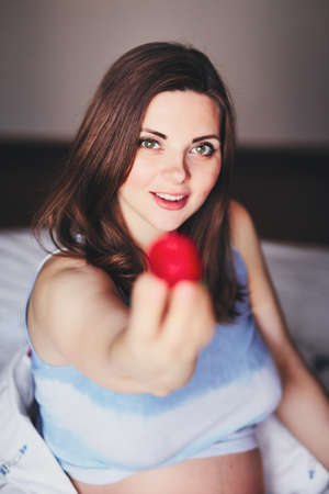 Attractive young pregnant woman sits on a bed and eats a ripe strawberry. Food for a happy pregnant girl, fruits and vegetables for a balanced diet future mom. Healthy food for a successful pregnancyの写真素材