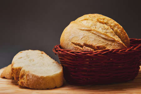Fresh bread made from wheat flour in a wicker basket made of dark wood lies on a wooden board and against a dark background. Lighting from above through the hole. Slices of freshly baked breadの写真素材