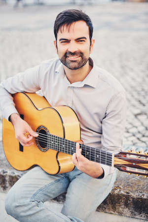 A cute stylish man with a beard sits on a concrete curb in the street and plays an acoustic guitar...