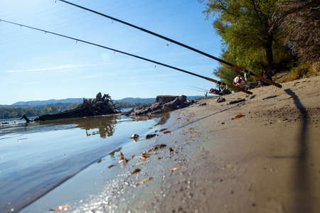 Two fishing rods are mounted on a rack located on a river sandy beach. The day is beautiful and...