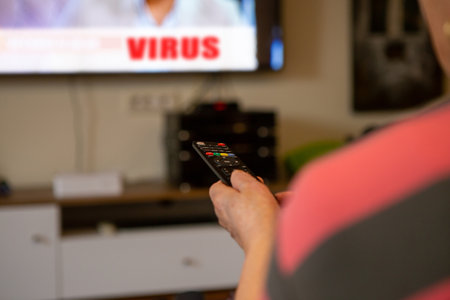 A senior woman sits in front of the television watching television. She holds a remote control in her hand that she points at the TV because she wants to change the channel. The focus is on the remote control.の写真素材