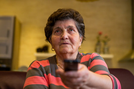 A senior woman sits in front of the television watching television. She holds a remote control in her hand that she points at the TV because she wants to change the channel. She is serious and concerned.の写真素材