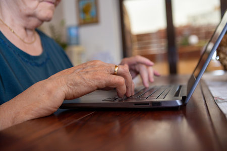 A senior woman with black short hair in a turquoise shirt is typing on a gray laptop keyboard. She is serious and thoughtful, she is in the dining room at the wooden table.の写真素材