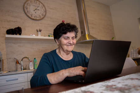 A senior woman with black short hair sits at a table in the kitchen and works on a laptop computer. She is looking for some new cooking recipes online and She is thoughtful and laughing.の写真素材