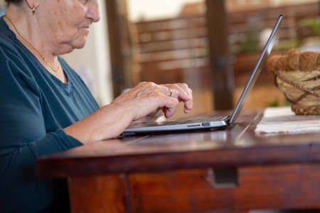 Senior woman wearing black short hair in a turquoise shirt writes on a gray laptop computer. She sits in the dining room at the wooden table and is thoughtful and serious.の写真素材