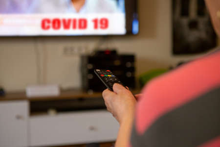 A senior woman sits in front of the television watching television. She holds a remote control in her hand that she points at the TV because she wants to change the channel. The focus is on the remote control.の写真素材