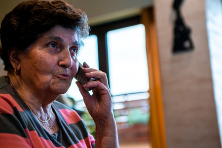 A senior woman with short black hair is sitting on a sofa in her home and talking on the phone. She looks very worried.の写真素材