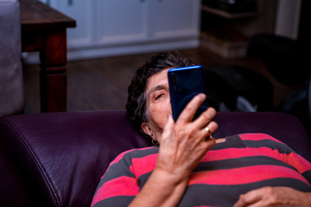 A senior woman with short black hair is lying on an armchair in her home and has a metallic blue smartphone in her hand. She is looking at something on the internet.の写真素材