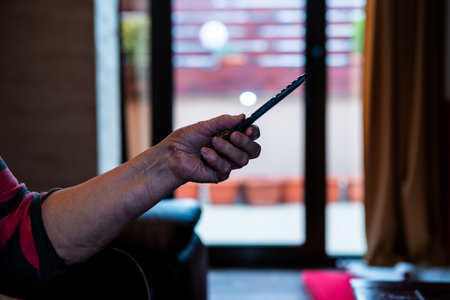 Selective focus on the TV remote control in the hand of a senior woman. She is sitting on the sofa in her home and watching television.の写真素材
