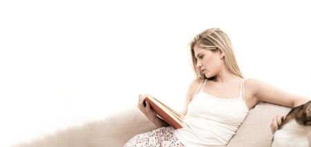 A young blonde sits on a sofa and reads a book. As she reads the book, she caresses her cat. She is very serious.の写真素材