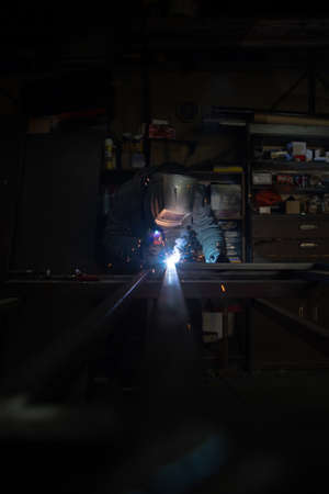 An adult male works in an old workshop. He holds a welding machine in his hand and welds the metal...