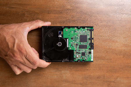 A man's hand holds a hard disk memory located on a wooden base. The hard drive shows electronics and a printed circuit board.の写真素材
