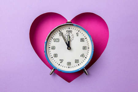 Old retro analog blue alarm clock in pink heart background on purple background. The clock starts from five minutes to twelve o'clock. Minimal concept of love, time and alarm.の写真素材
