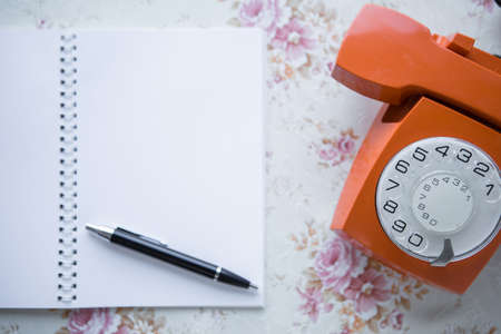 Selective focus on old analog orange phone. Next to the phone are a notebook and a pen. Copy space....