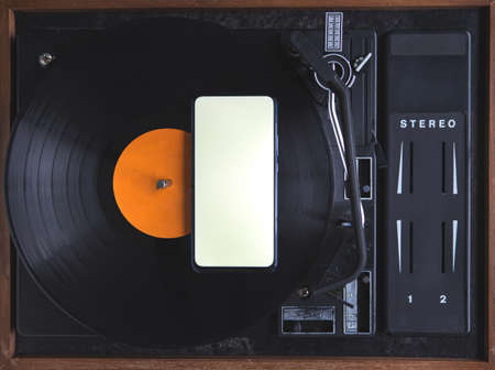The top view of the new modern smartphone is on the music record on the turntable. The phone has a white screen. Music. Application. Copy space. Old and modern.の写真素材