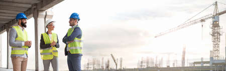A beautiful female engineer with her handsome colleagues on a construction site.And behind in the distance you can see the construction site and construction cranesの写真素材