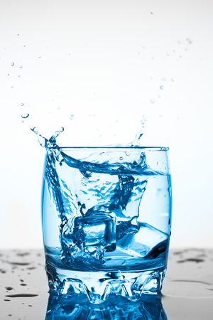 water splash in a glass with a piece of ice on a white background, a piece of ice falling into glass Cup of water, splashes of water flying in all directionsの写真素材