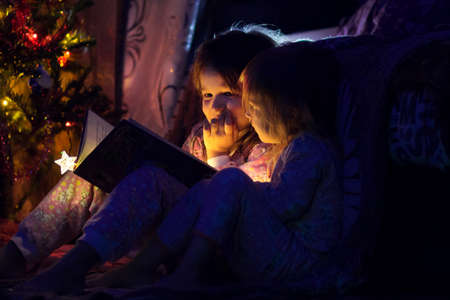 Two sisters in pajamas read the magic book near a New Year treeの写真素材
