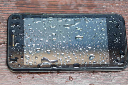 Close up of phone which is filled in with drops which lies on a brown wooden table.の写真素材
