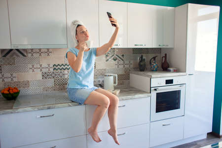 Woman with a towel on her head in her kitchen having breakfast and looking at the phoneの写真素材