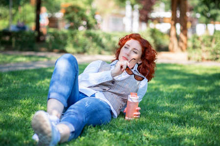 A cute red-haired girl lies in the park on the grass and thinks or dreams about something. Woman walking in a summer park with a bottle of waterの写真素材