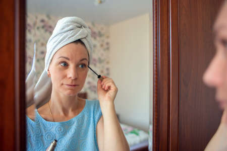 A girl with a towel on her head paints her eyelashes with mascara. The woman is going to work. Morning toilet.の写真素材