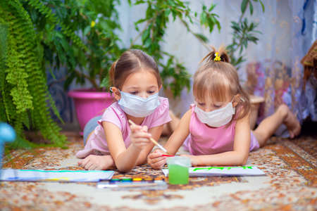 Two girls in protective medical masks paint with paints lying on the floor at home or in kindergarten. Children in self-isolation or quarantineの写真素材