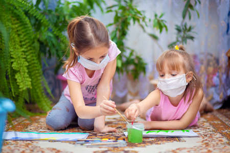 Two girls in protective medical masks paint with paints lying on the floor at home or in kindergarten. Children in self-isolation or quarantineの写真素材