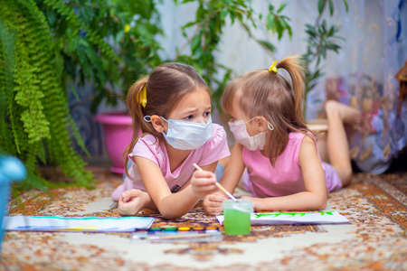 Two girls in protective medical masks paint with paints lying on the floor at home or in kindergarten. Children in self-isolation or quarantineの写真素材