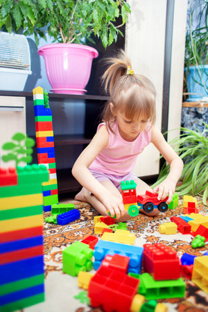 A little girl sits on the floor at home and plays a multi-colored plastic constructor. A child playing a toy trainの写真素材