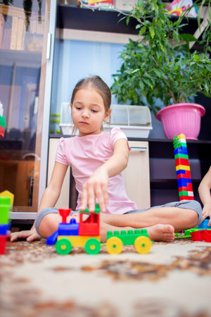 A little girl sits on the floor at home and plays a multi-colored plastic constructor. A child playing a toy trainの写真素材