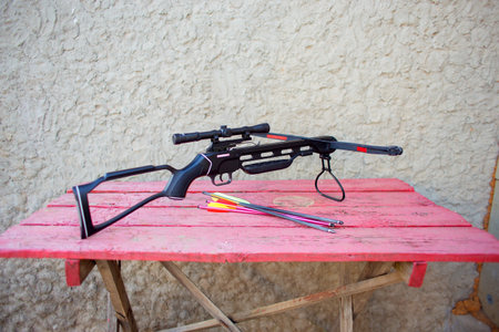 A modern crossbow with arrows and a telescopic sight lies on a wooden table in the street. Preparing...
