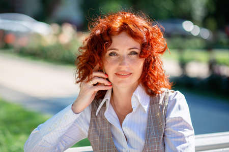 Beautiful red-haired woman in the summer sits on a bench in the park and uses the phoneの写真素材