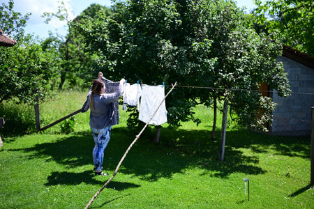 Woman Hanging Laundry Outdoors on a Sunny Day in a Serene Countryside Garden with Lush Greenery and...