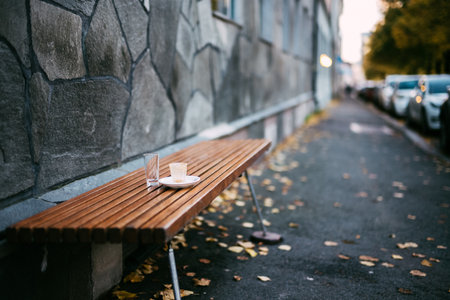 Wooden Park Bench with Coffee Cup in Autumn Urban Street Lined with Cars and Stone Wall Background