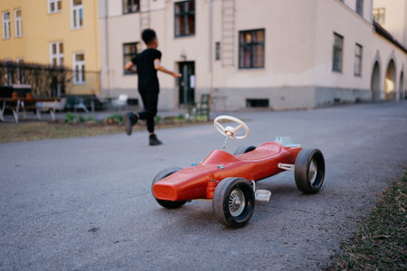 Cheerful Boy Running Past a Red Toy Car on a Sunny Day in a Scenic Urban Street Setting