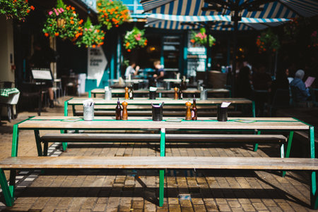 Charming Outdoor Cafe Scene with Empty Picnic Tables Under Bright Sunlit Striped Awnings and Hanging...