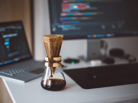 Modern Home Office Setup with Chemex Coffee Brewer on Desk and Coding in Background