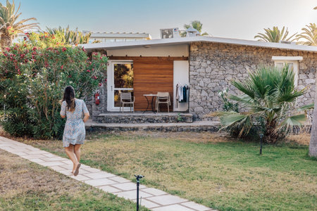 Charming Stone Bungalow with Vibrant Garden and Woman Strolling on Serene Pathway in Summer