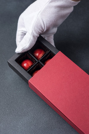 Hand in white glove holding red box with chocolates on black backgroundの写真素材