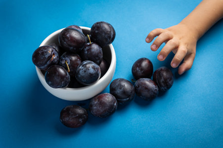 A child's hand reaches for a bowl of plums on a blue backgroundの写真素材