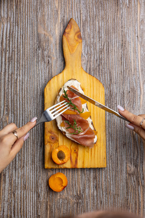 Top view of woman's hands holding fork and knife with fresh prosciutto on wooden boardの写真素材