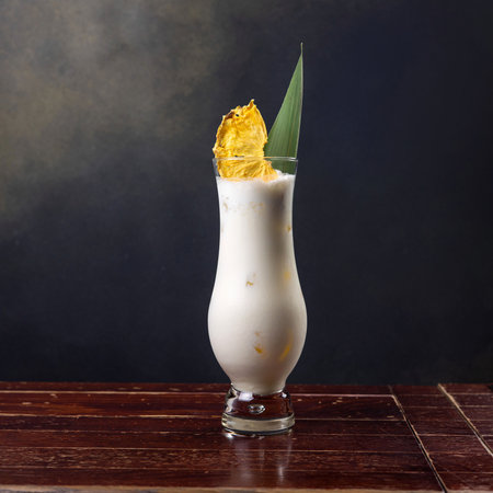 Beverage in a tall glass on a dark background. Drink decorated with a slice of pineapple and a green...