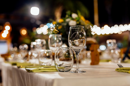 Romantically set dinner table with wine glasses and floral centerpiece, perfect for a wedding, elegant party, or outdoor evening celebration. Warm lights and soft bokeh create an intimate and luxurious atmosphere.の写真素材